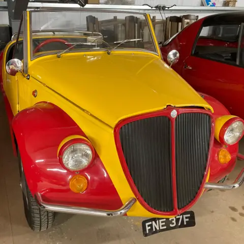 Mobil kuno dengan dua tempat duduk dengan bagian atas terbuka dan dicat cerah dengan warna merah dan kuning.