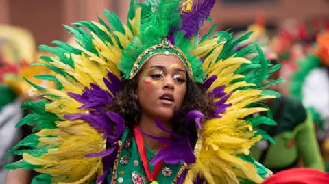 Andrew Aitch/Manchester Carnival A young woman wears a large head-dress of purple, yellow and green fake feathers as she dances in a parade. She wears red, green and yellow sparkles around her eyes.
