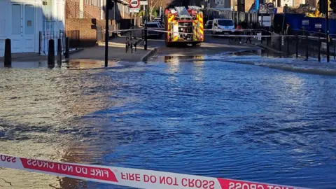 A view of flood water behind a cordon. There is a fire engine parked on the opposite side of the water. 