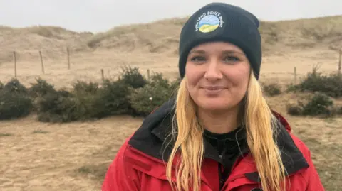 Amy Pennington, who is wearing a red coat and hat with the words Fylde Sand Dunes Project, is standing in front of rows of old Christmas trees on the sand dunes. 
