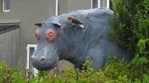 Malcolm Curley A hippo sculpture casually leans over a garden hedge with a bird sitting on its neck. There's a house in the background.