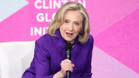 Hillary Clinton, wearing a purple suit, steps forward and speaks into a microphone on stage at the event 