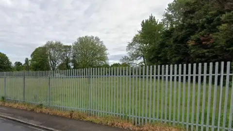 An image of a field with grey fencing. There are trees in the background.