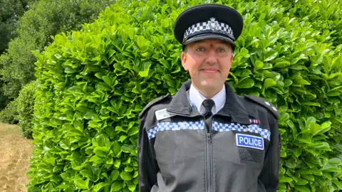 A police officer in a uniform stands in front of a large green bush.