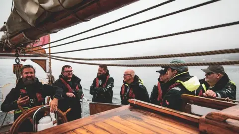 Turn to Starboard Seven men in heavy weather gear and life belts are standing near the wheelhouse of a wooden ship with the sails reefed above them. Some of are smiling, some are looking pensive as they listen. 