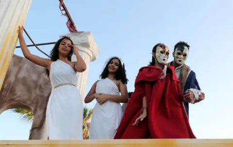 MOHAMED MESSARA / EPA People dressed in white togas and Venetian ball attire stand on a moving float that has Greco-Roman columns in the background.
