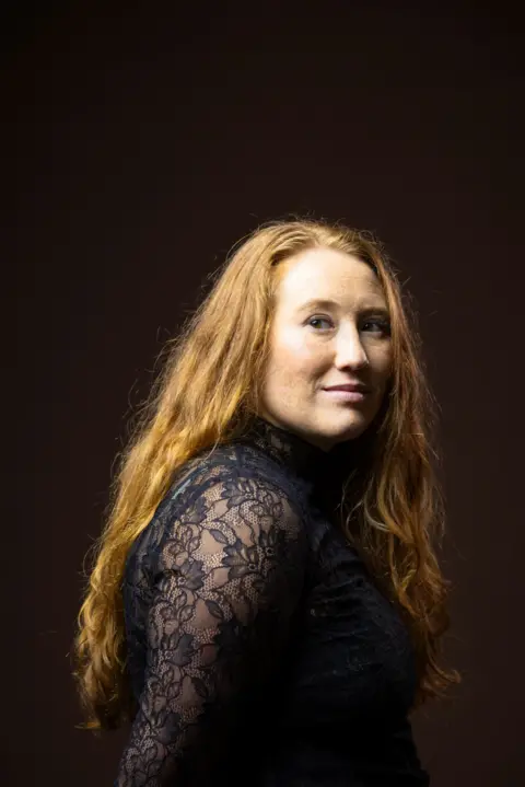 Kieran Dodds Studio portrait of a person with long, wavy hair wearing a black high-neck lace top with sheer floral-patterned sleeves. The person is turned slightly sideways while looking toward the camera. Warm studio lighting highlights the hair texture and intricate lace details against a dark brown background.