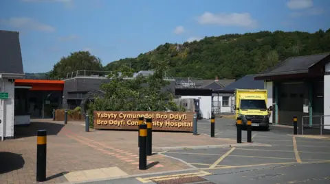 The picture shows part of the Bro Dyfi Community Hopsital site. There is a sign with the hospital name on. Next to it is an ambulance, yellow and green in colour. There are bollards surrounding the sign and ambulance. The photo is taken on a sunny day, with blue skies at the top of the picture.