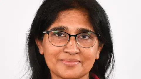 Luton Borough Council Studio-style head-and-shoulders portrait of Rumi Choudhury with long dark hair, wearing glasses and a red jacket, against a plain light background.