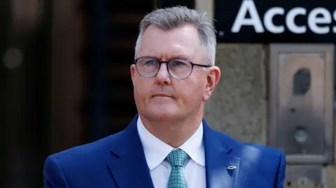 Sir Jeffrey Donaldson has short, cropped grey hair. He is wearing black-rimmed glasses and a blue suit, white shirt and a green patterned tie