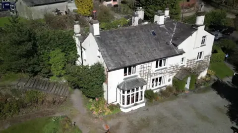 An aerial view of the large, white house on a sunny day. There are trees next to it as well as what looks like a farm.