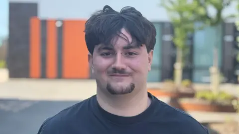 Jamie is smiling at the camera. He has dark hair with a floppy fringe, a goatee beard and studs in his nose. He is wearing a black T-shirt and just his head and shoulders are in shot - with the sixth form college and trees behind him.