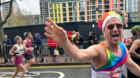 Family photo A man running a race is wearing a rainbow coloured outfit and hat, along with sunglasses. He is waving to the camera.
