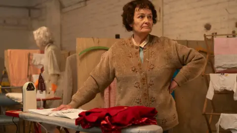 BBC/World Productions Ella Ramsden, played by Estrid Barton, standing by an ironing board with items of clothing; she's an older woman in a brown cardigan
