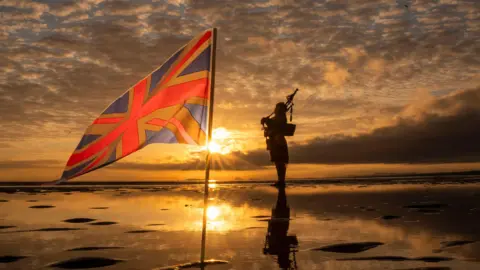 A piper stands against the sun in an orange cloudy sky beside a union flag in the wet sand.
