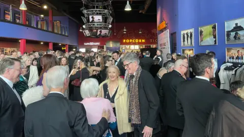 The event in the cinema is packed with smartly dressed people. There are photos from the TV shoot on the wall.