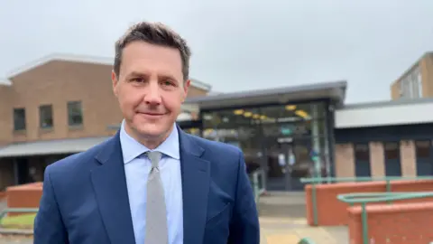 Head teacher Stuart Shelton in front of St Peter’s Catholic School, a brick building with a glass reception area. He is wearing a light blue shirt, a tie and a blue suit.