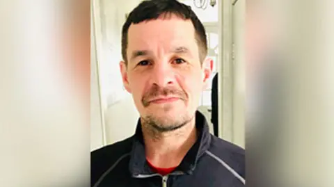 Police handout Craig Foy with brown hair and brown stubble wearing a navy zip up jumper standing in a hallway with a white front door behind him. 
