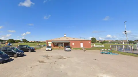 We can see a car park in front of a pavilion, with part of the playground to the left and the multi-use games area on the right. It is a sunny day with a bright blue sky.