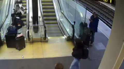 GTR Passenger luggage tumbles down an escalator at a railway station platform, with two people also on the escalator.