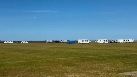 A row of white caravans have parked along the edge of a football pitch. There are tyre tracks over the pitch leading to them.