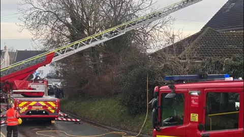 Two red fire engines on the road- with an extended metal ladder from one of them- and crews standing on the road- wearing orange jackets 