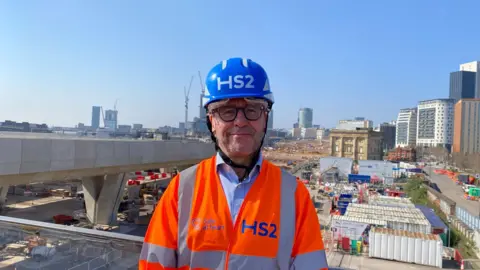 Alexander Brock A man in a blue hard hat with 'HS2' on it, and an orange hi-viz jacket also saying HS2. He stands with the HS2 construction site in Birmingham behind him, as the bridge is made.
