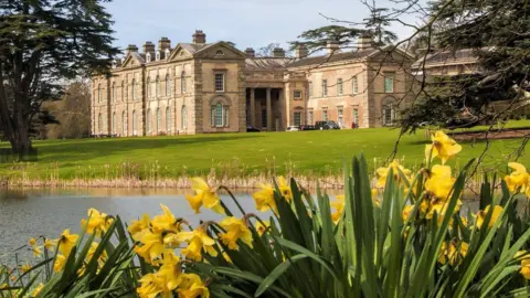 Compton Verney A large historic country house stands beside a lake, with bright yellow daffodils in the foreground and manicured lawns and mature trees surrounding the building on a sunny day.