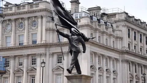 A grey statue of a man wearing a suit, marching forward, carrying a flag that covers his face
