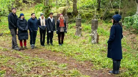 BBC/Studio Lambert Six contestants from the Traitors stand in a wooded area surrounded by greenery and in front of some hanging baskets. They are all wearing jackets and warm outerwear. Claudia Winkleman is standing in front of them.