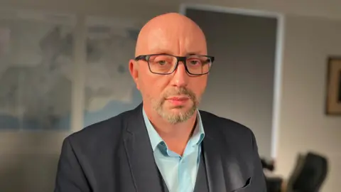 BBC David Hinton in a light blue shirt and dark jacket. he is looking at the camera and has a stern expression.