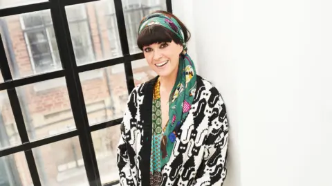 BBC A smiling woman, Dawn O'Porter, with dark hair, heavy eye makeup, and a fringe leans against cast-iron window with a brick building in the background. She has a teal headscarf with yellow and purple patterns, and wears a black and white oversized cardigan with a tessellated pattern of cats on it.