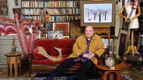 Desmond Morris at home. This looks like a busy sitting room with Morris sitting on a red couch surrounded by books and his art works.