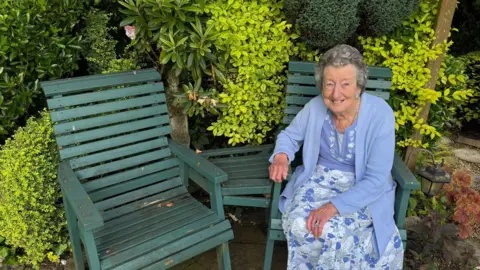Ann Lush Ann Lush sat in her garden on a green wooden chair. Next to her is an empty matching chair. She is an elderly woman wearing a blue and white floral dress, with a matching blue cardigan. She is smiling at the camera
