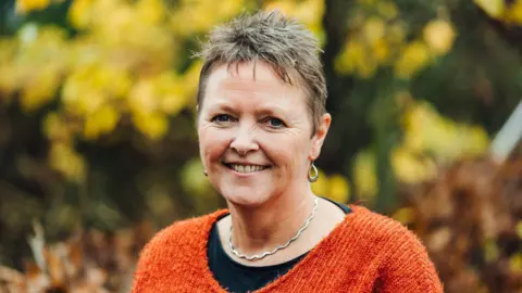 A woman with short grey hair smiles at the camera in an outdoor portrait. She is wearing a bright orange knitted jumper over a dark top, with a silver necklace and silver hoop earrings. The background is softly blurred with autumn foliage in shades of gold, green and brown.