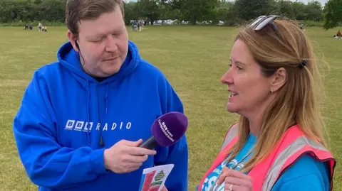 A BBC Radio microphone is held out during an interview at an outdoor festival, with two people standing on grass and open parkland visible in the background.