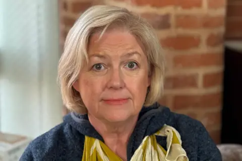 A close-up of a woman with shoulder-length blonde hair who is looking at the camera. She wears a dark fleece and a yellow floral scarf.