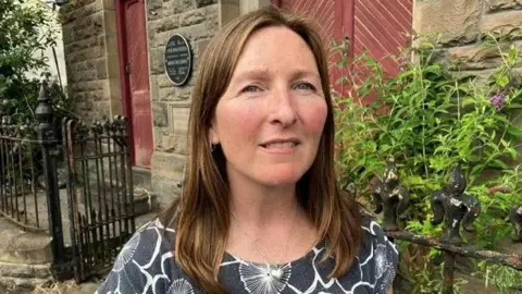 Rhian Hopkins outside the chapel in grey top with white pattern of flowers. She has long light brown hair and wears a silver heart-shaped necklace