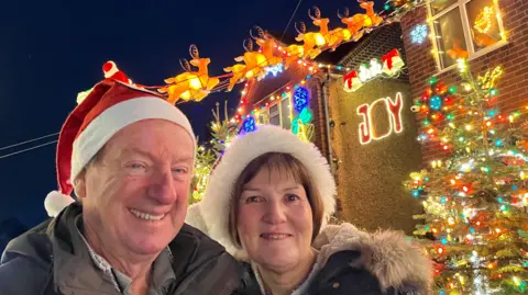 Chris and Christine stand outside a house wearing Santa hats. Behind them is a lit up sign saying "Joy", as well as a Christmas tree and reindeer decorations