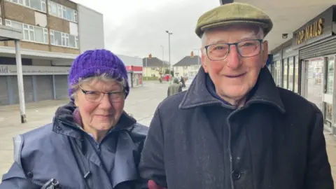 Barbara Dawson is smiling. She has grey hair and glasses and is wearing a purple wool hat and a navy blue coat. She is standing next to John Dawson who is taller. He is wearing a flat green cap and is also smiling. He is also wearing glasses and a navy blue coat. There are houses behind them and shops with people walking around