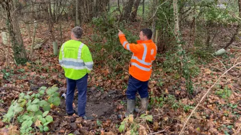 An Environment Agency and Welsh Water worker stand in woodland by the river. they both have fluorescent jackets on and wellies.
