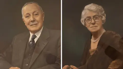 The Mitchell family Portraits of Fred Mitchell (right) and his wife Clarrie (left). He has grey hair and a grey moustache, and is wearing a grey waistcoat and jacket, white shirt and blue tie. She has short, grey hair and is wearing a pearl necklace, glasses and fur jacket