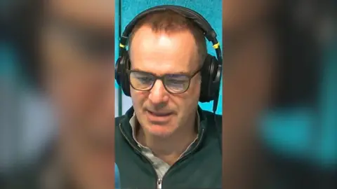 Martin Lewis has short dark hair and is wearing radio headphones, dark rimmed glasses and a green quarter-zip.