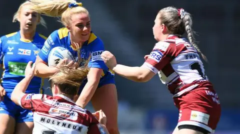 Lucy Murray of Leeds Rhinos is challenged by Victoria Molyneux and Rachel Thompson of Wigan Warriors during the Betfred Women's Challenge Cup Semi-Final match between Wigan Warriors and Leeds Rhinos at Totally Wicked Stadium on May 19, 2024 in St Helens, England