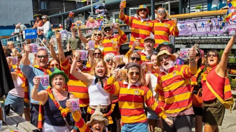 A large group of people are gathered outside a harbourside pub, many wearing matching red and yellow striped tops while holding up drinks and plastic cups towards the camera. Colourful maritime flags hang above the crowd, and wooden benches, buildings and festival signage can be seen in the busy, sunny background.