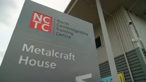 Andy Meeson/BBC Looking up at a rectangular sign standing outside a building. The sign is grey and says "North Cambridgeshire Training Centre" at the top, next to a red square with the letters NCTC inside, and "Metalcraft House" lower down. Behind the sign is an industrial-looking building, which has metal walls.