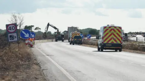 An empty dual carriageway road with work vehicles parked. There is a mechanical claw working on one side of the road next to a low barrier. On either side of the road you can see low vegetation and some trees. 