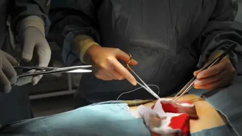 A stock image of surgeons carrying out an operation on a patient. The image is a close-up of hands holding tools to stitch up a wound, with gauze with some blood around it.