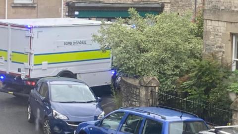 A white bomb disposal vehicle outside a home, next to two blue cars. 
