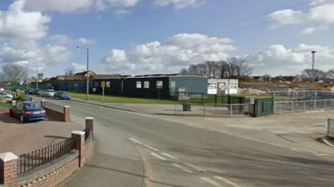 The image shows a quiet suburban street on a bright day with a partly cloudy sky. In the foreground on the left, there is a red-brick house with a grey tiled roof, bordered by a low brick wall and black metal fencing. A paved driveway or walkway runs alongside the house, and a small brick wall separates the property from the road.